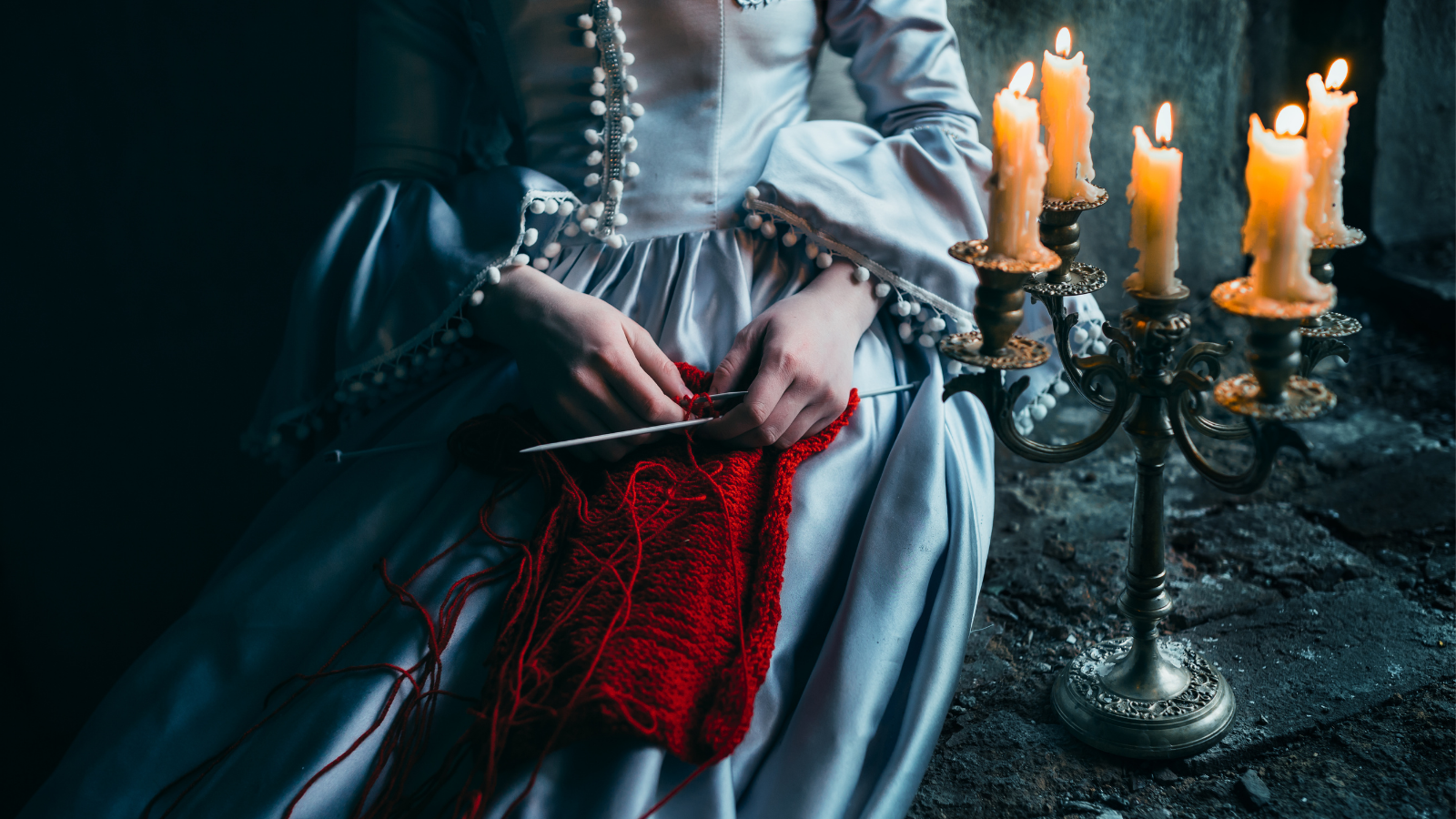 Person in a dress sitting beside a candelabra while knitting.